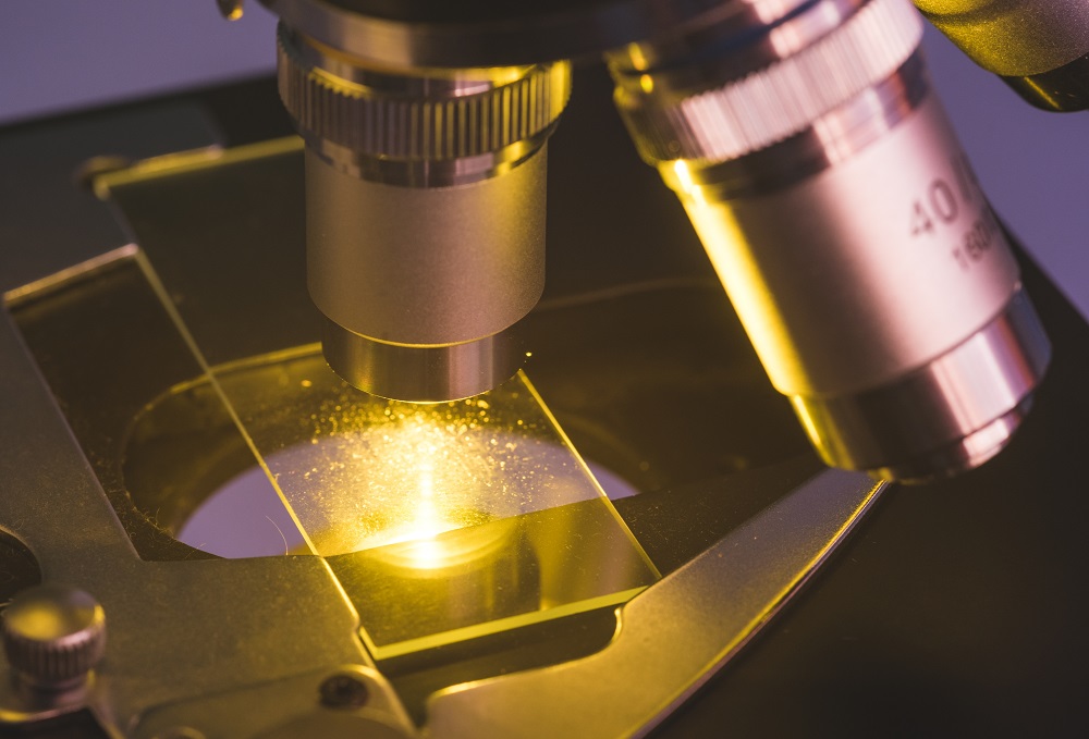 Close-up view of a microscope lens focusing on a glass slide with illuminated particles or specimens visible under the light.